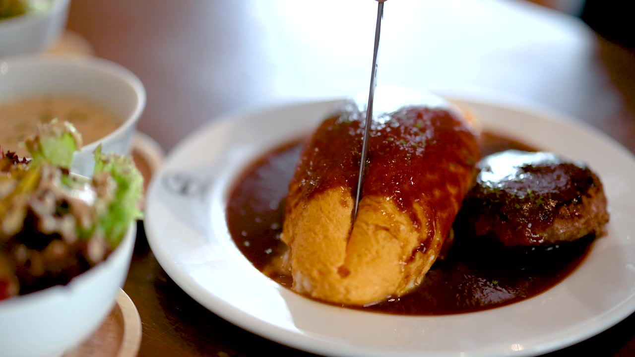 Knife slices omurice with demi-glace beside hamburg steak, natural light, shallow depth of field