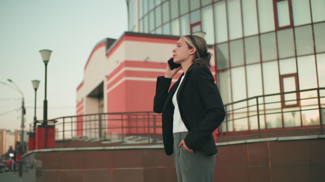 Professional lady in business attire talking on phone with frustrated expression, pacing near fence in urban environment, standing against backdrop of red modern building with reflective glass panels