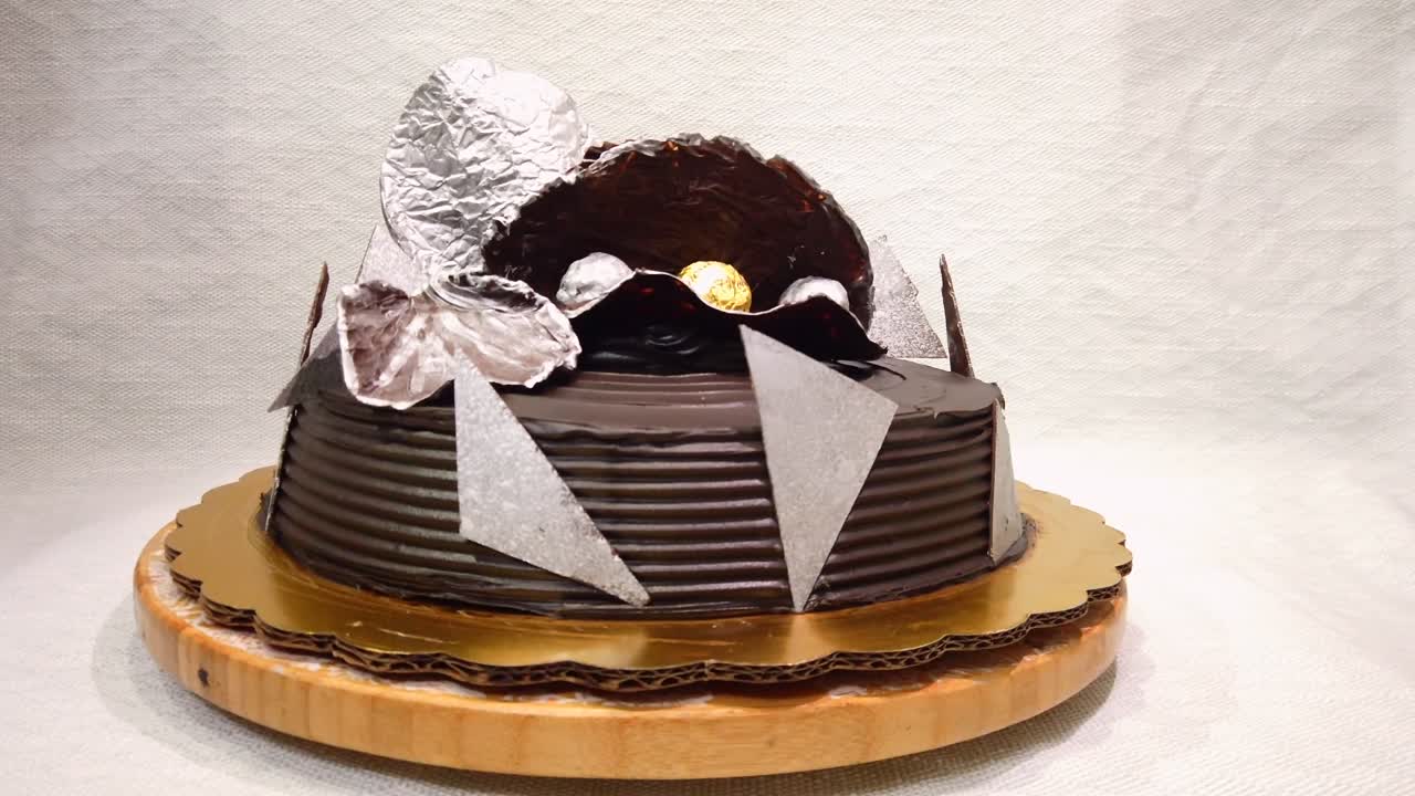 Torta al cioccolato con glassa testurizzata e scaglie di cioccolato decorative che ruota su una base mentre una mano la gira delicatamente. Primo piano.