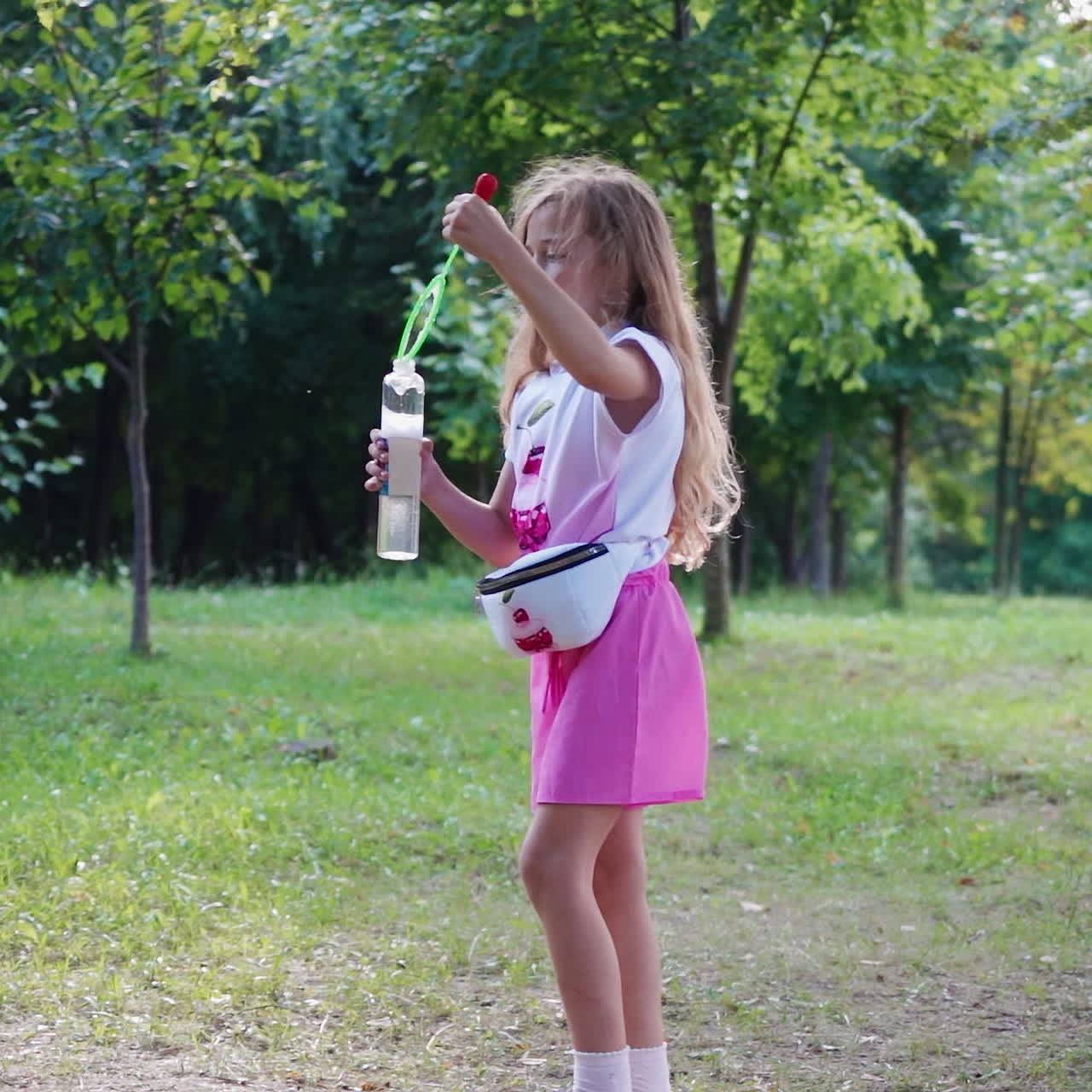 Side view of girl with bubble blower outdoors. Cute little girl in pink suit with long hair blowing soap bubbles in summer. Summer vacation concept.