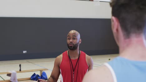 Entrenador-De-Baloncesto-Masculino-Diverso-Con-Portapapeles-Instruyendo-Al-Equipo-En-La-Cancha-Cubierta,-En-Cámara-Lenta