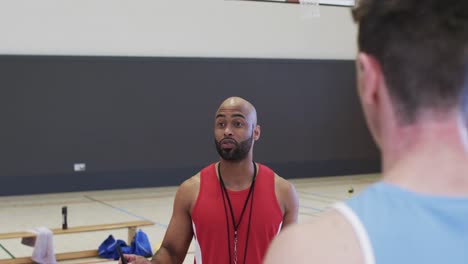 diverse male basketball coach with clipboard instructing team at indoor court, in slow motion