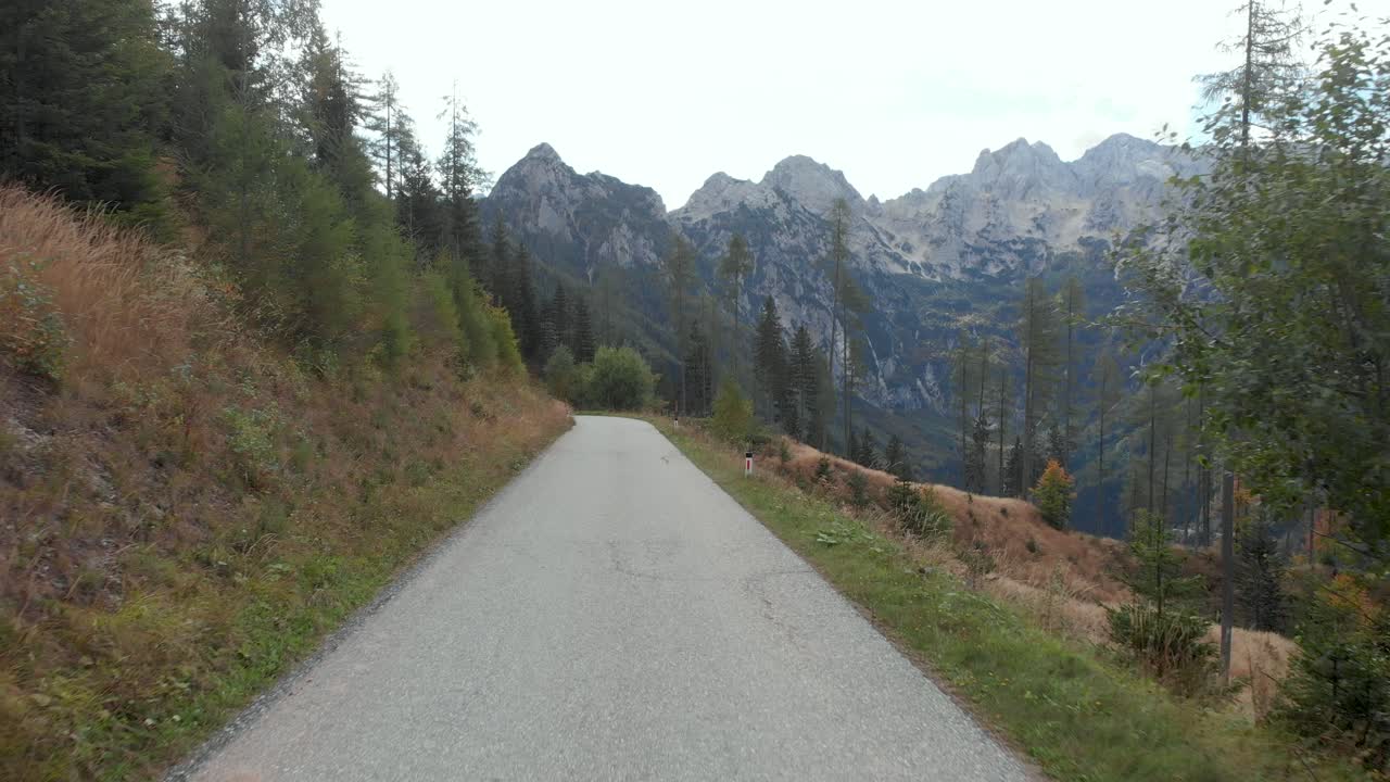 Drone flying along the road as it lifts up showing the tight road curving around the mountains with a car driving around it. Jezesko Valley in Slovenia in Autumn season aerial view