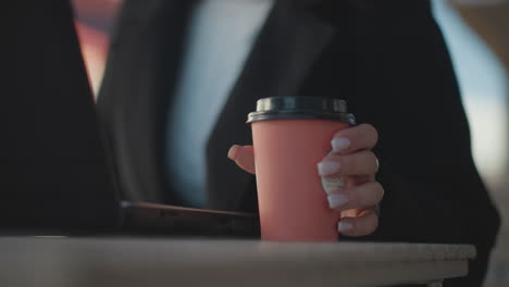 close-up of laptop on table with hand in black coat, polished nails, dropping coffee cup onto table surface, someone sitting down, blurred background with outdoor restaurant setting