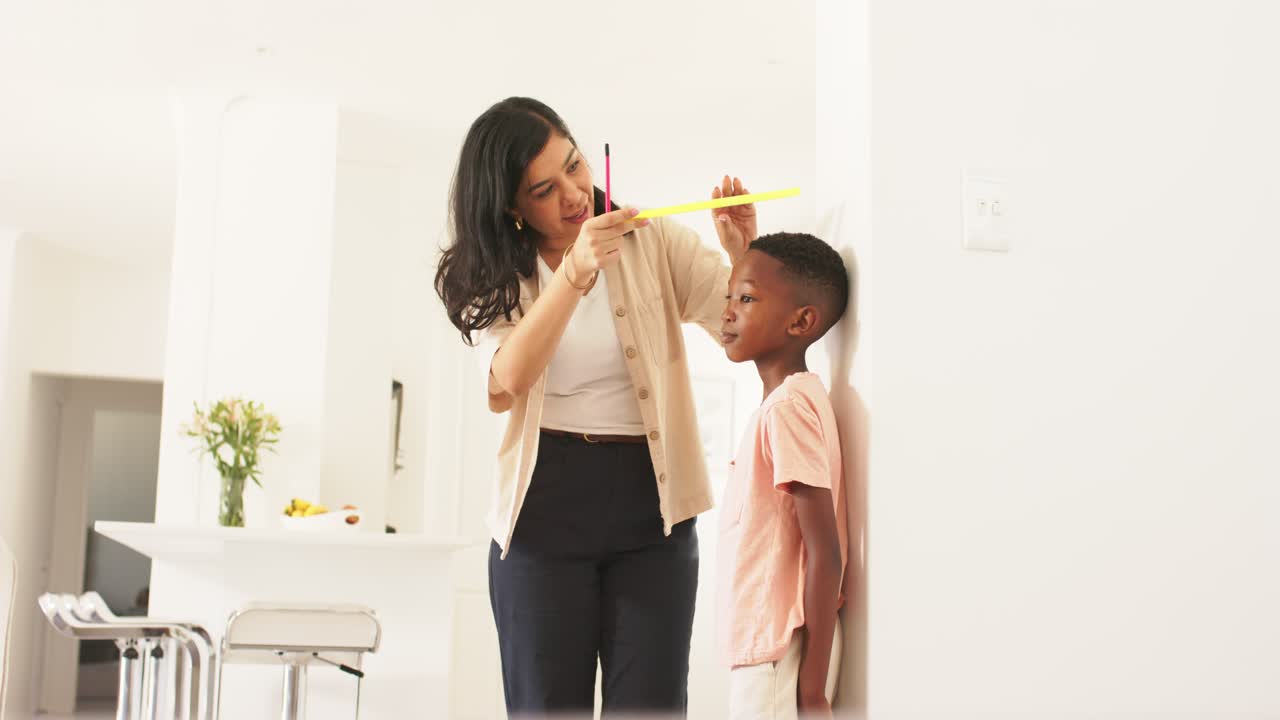 Diverse mom measuring African American boy against white wall using yellow tape, recording height