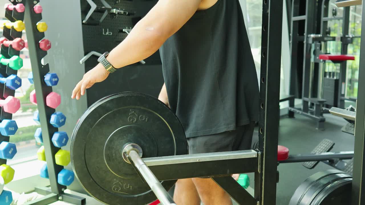 Two individuals in a gym setting prepare a barbell for a workout. Bright lighting highlights the equipment and focused atmosphere