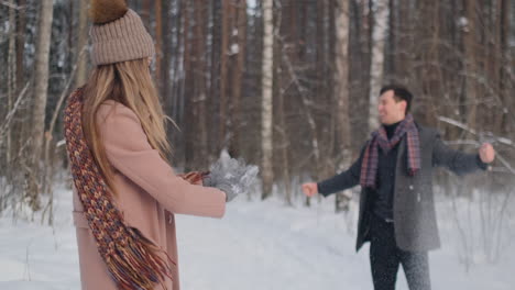 Ein-Junger-Mann-Und-Eine-Frau-Im-Mantel-Amüsieren-Sich-Und-Spielen-In-Zeitlupe-Mit-Schnee-In-Einem-Winterwald.-Glück-Und-Lächeln-Auf-Den-Gesichtern