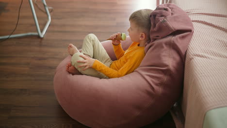 young boy sitting relaxed on large pink beanbag licking mint chocolate ice cream cone while looking toward background where metal stand is visible on wooden floor