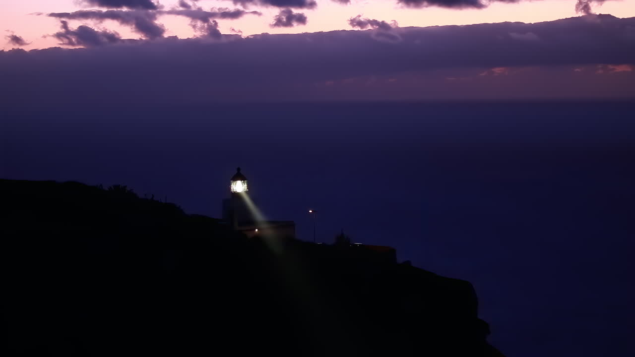 A lighthouse stands proudly on a cliff as the sun sets, illuminating the surroundings with its bright light. The skies are filled with soft colors as night approaches