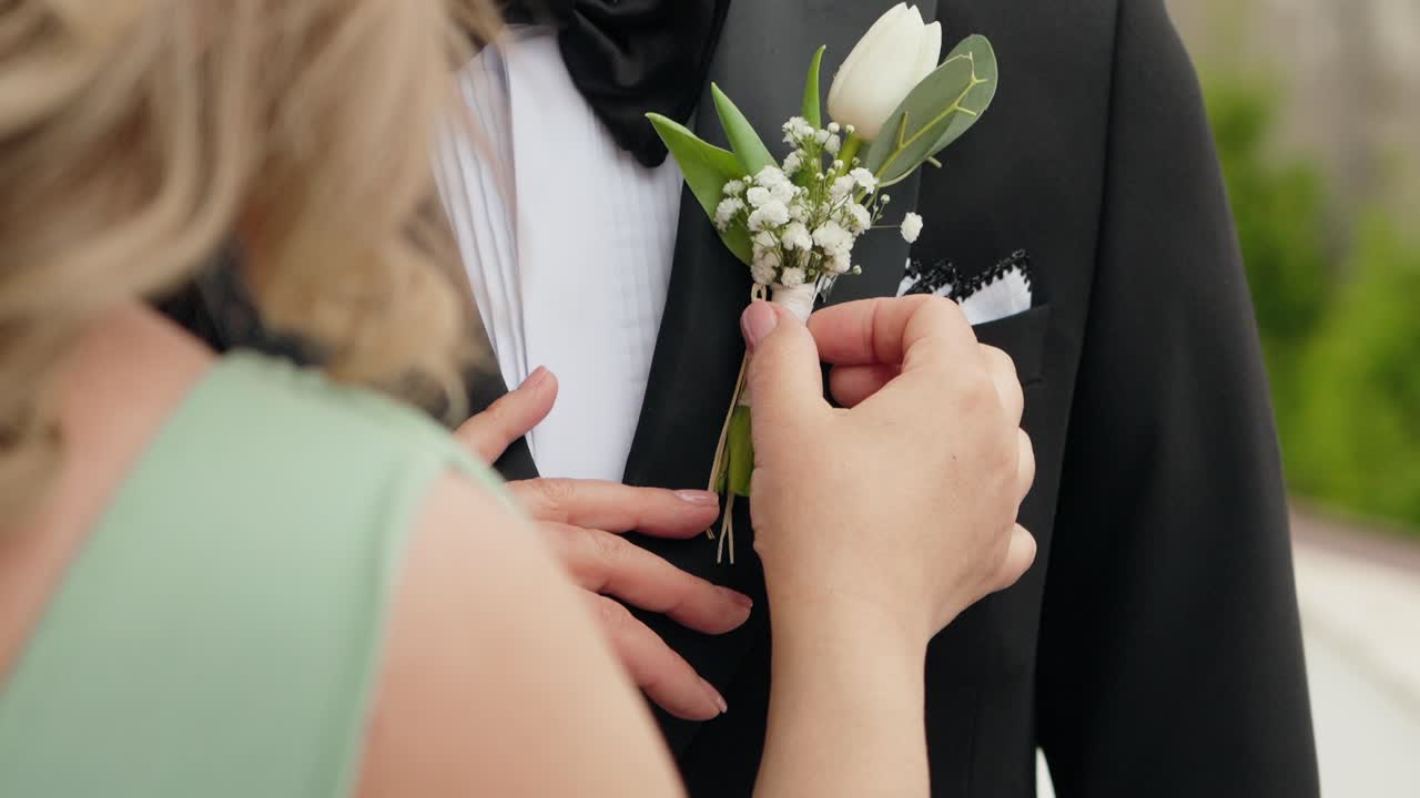 woman pins white boutonniere on groom’s black tuxedo with gentle focus on hands and flower