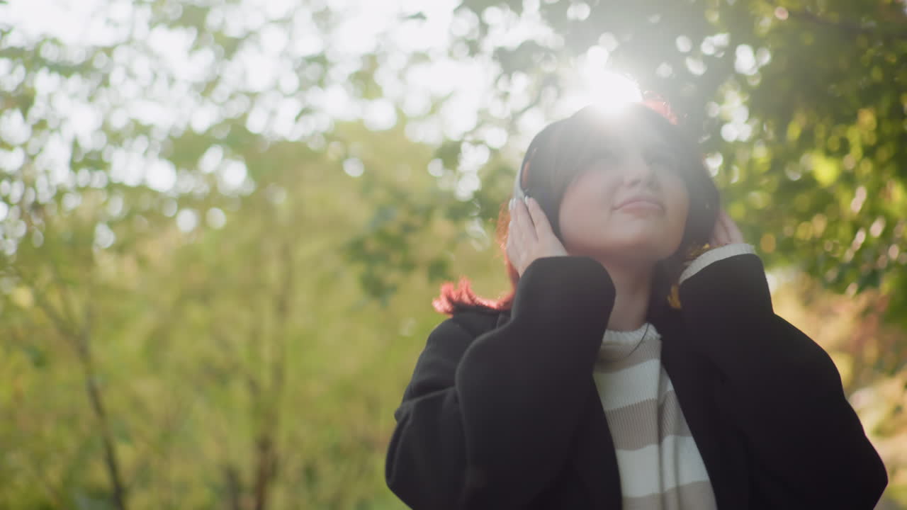 Medium view of free spirited lady wearing headphones, spinning round then strolling through autumn park under soft sunlight, relaxed mood with yellow leaves on shoulder, playful movement