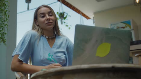una chica en un vestido azul remotamente en línea trabajando detrás de una computadora portátil y mirando a la pantalla el patio trasero con plantas verdes