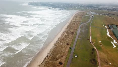 Sea-waves-crashing-on-long-beach-and-St-Andrews-city-in-background,-Scotland