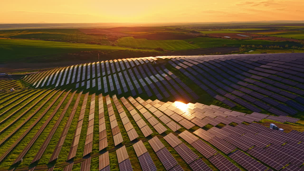 Solpaneler glimter under solnedgangshimlen ved en vedvarende energipark på grønne marker med fjerne bakker og skyer.