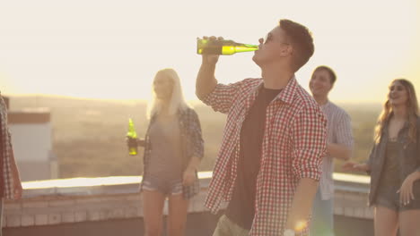 a young man in trendy glasses drinks beer and moves in a dance at a party with his friends on the roof.