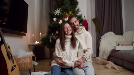 Portrait-of-a-happy-couple,-a-brunette-guy-with-stubble-in-a-white-sweater-and-a-brunette-girl-in-a-White-sweater-sitting-on-the-floor-near-their-Christmas-tree-and-their-cream-colored-cat-in-a-cozy-house-decorated-for-a-Christmas-atmosphere-in-winter