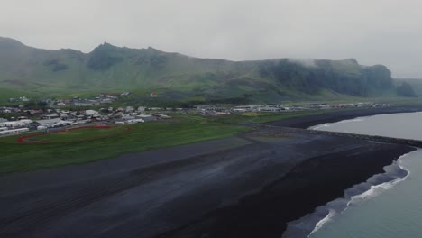 Orbiting-aerial-drone-shot-of-beautiful-blue-waves-crashing-on-a-black-sand-beach-in-front-of-Vik,-Iceland-and-huge-rock-formations-and-mountains-in-the-distance