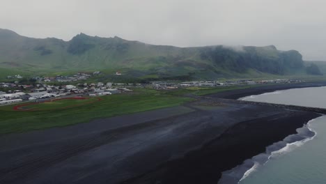 toma aérea en órbita de drones de hermosas olas azules rompiendo en una playa de arena negra frente a vik, islandia y enormes formaciones rocosas y montañas en la distancia