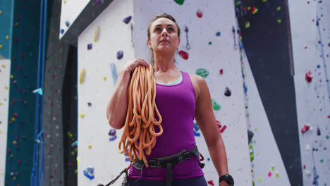 Mujer-Caucásica-Con-Una-Cuerda-Sobre-Su-Hombro-Preparándose-Para-Escalar-En-El-Muro-De-Escalada-Interior