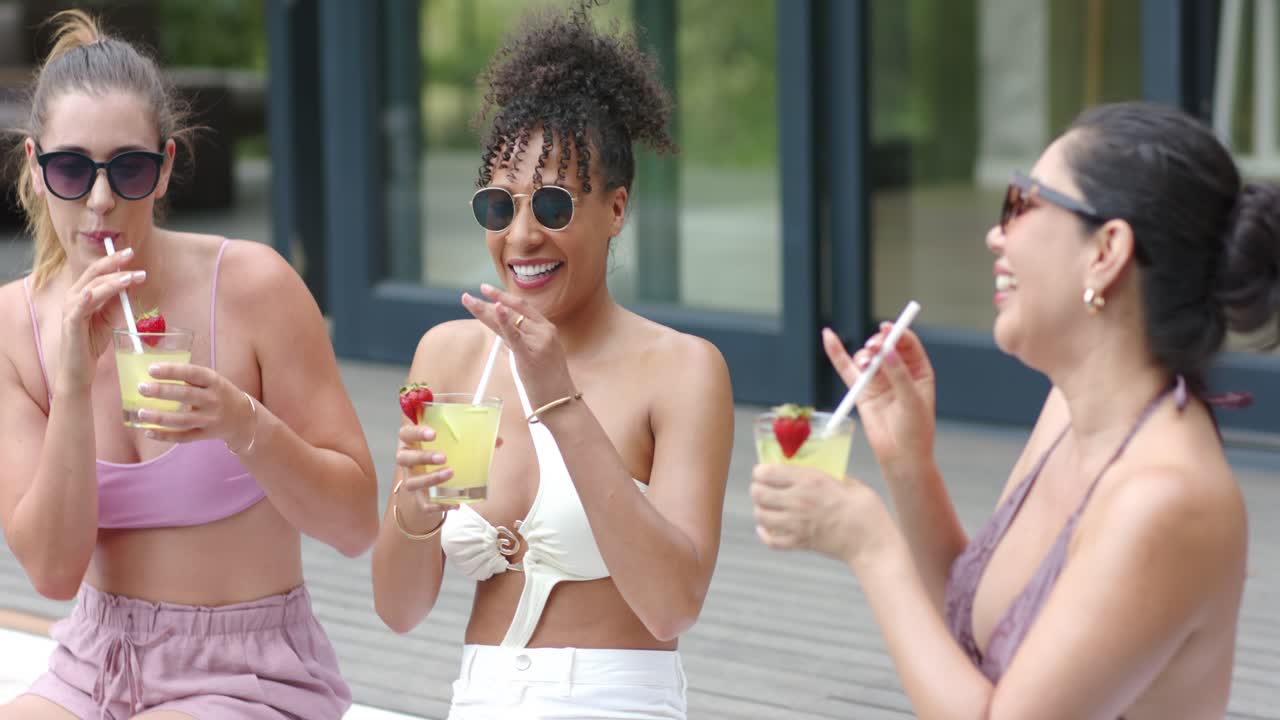 Diverse female friends starting toast, clinking and sipping cocktails by pool wearing sunglasses