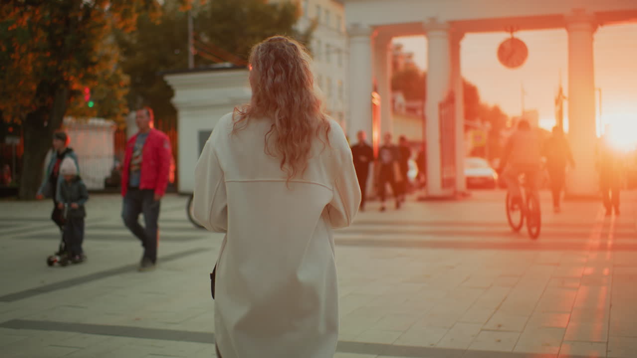 back view of girl in long white coat strolling through city walkway during golden sunset as glowing sunlight creates radiant atmosphere with people walking around
