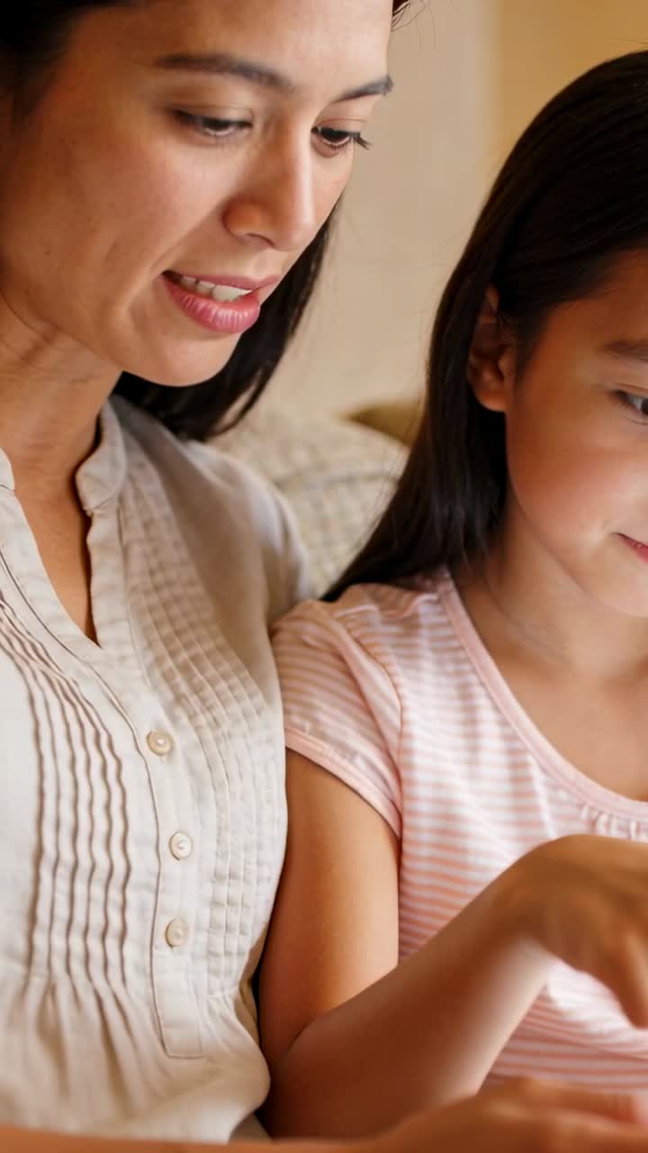 縦型動画：ソファでデバイスを共有する母娘。ブラウス姿の母親が指導する中、子供がタップしている。