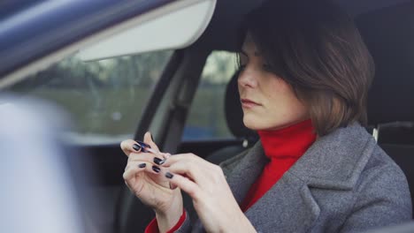 Business-woman-in-grey-coat-and-red-turtleneck-applying-makeup-in-the-car