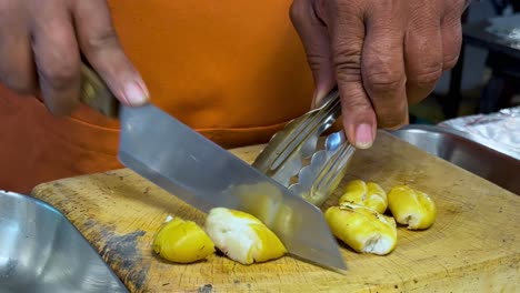 close-up of hands skillfully slicing yellow fruit using a knife and tongs on a wooden board.