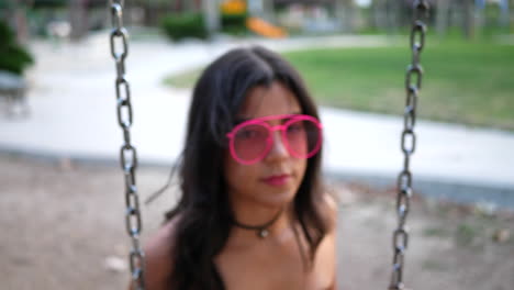 una hermosa modelo de moda hipster con ropa vintage y gafas de sol rosas retro que se balancean en un patio de recreo y se divierten a cámara lenta