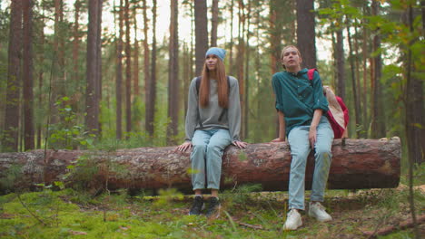 two tired hikers sit on fallen tree in lush forest, one with blue bandana lifts leg happily, appearing relaxed, while companion in green shirt rests hand on tree, with tall trees around