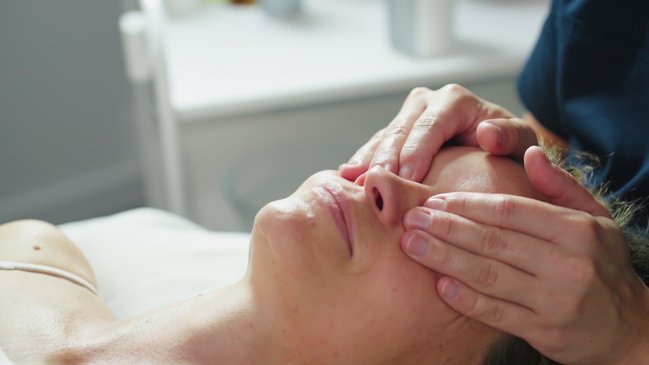 High angle close up of cosmetologist placing her hands on lady lying on massage bed gently massaging around eye area with soothing strokes under soft spa lighting for calming eye treatment session