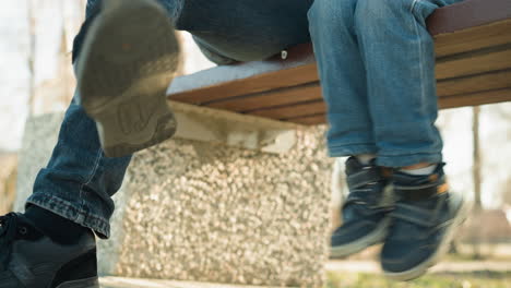 close-up of an adult and a child sitting closely together on a bench, the child playfully dangles his legs, gently tapping the adult s crossed legs, with a blurred background of trees