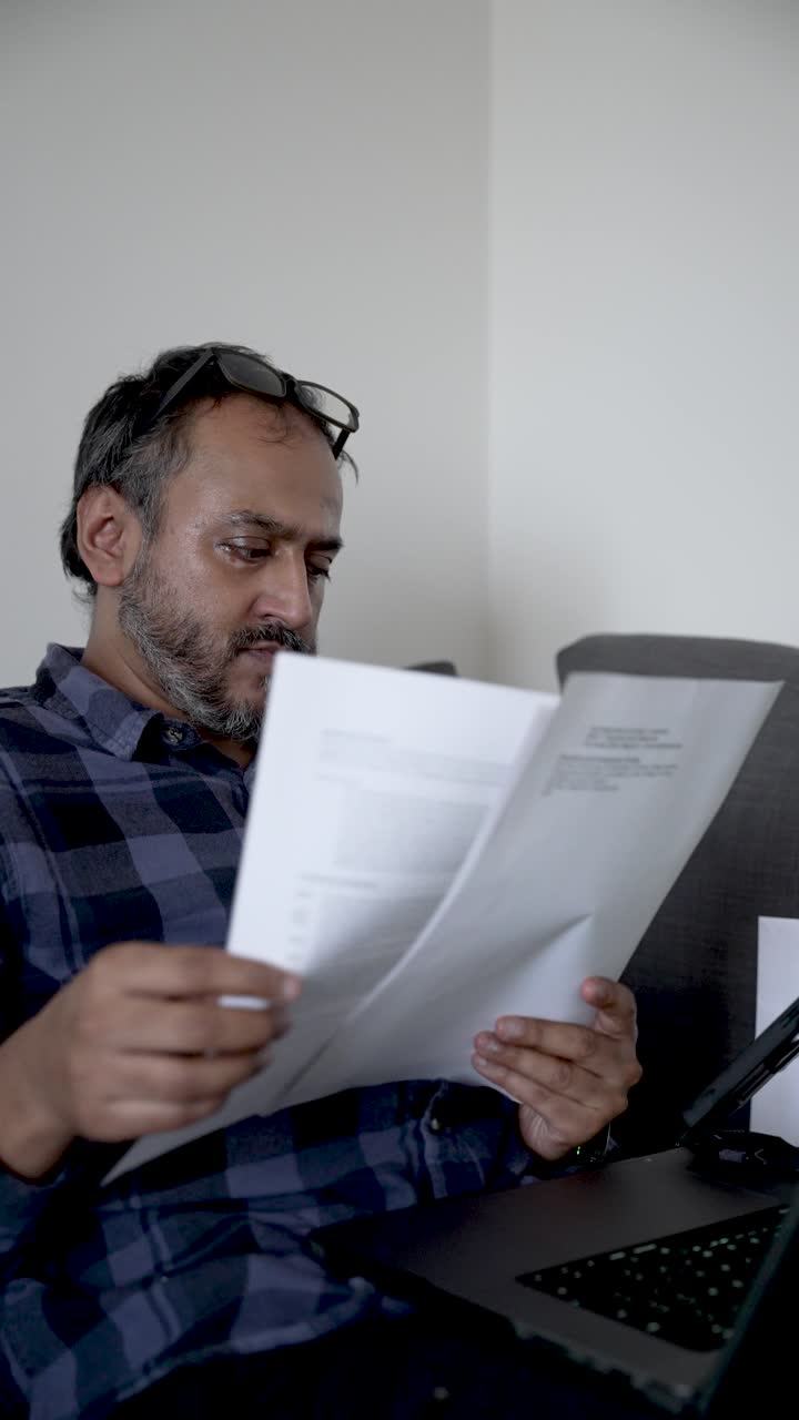 Trabalho remoto e auditoria de documentos: um homem sul-asiático com óculos na cabeça verifica documentos. Vídeo vertical.
