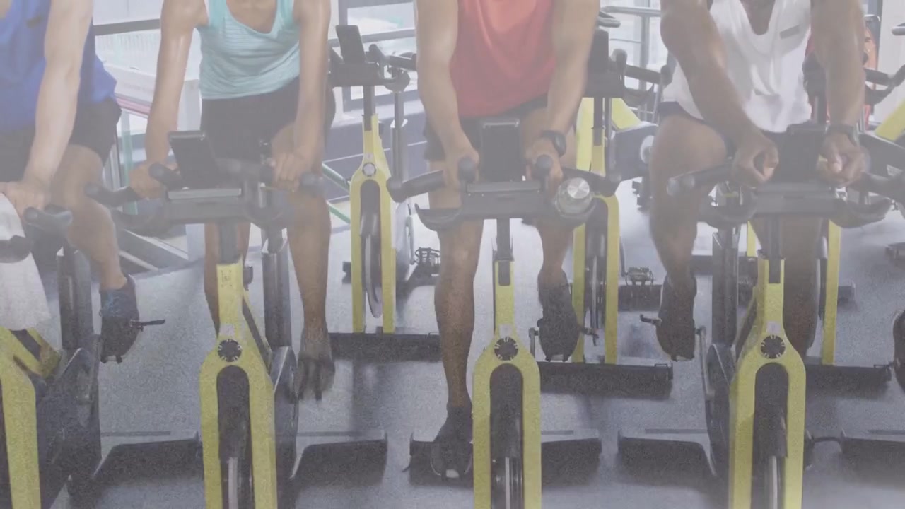 Ciclistas en clase de spinning siguiendo las indicaciones, pedaleando, ajustando los agarres, números de la consola actualizándose.