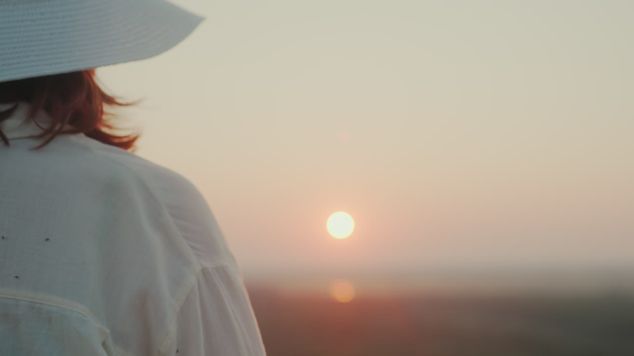 Close up rear view of woman in wide brim hat turning toward camera with warm smile while glowing sunset illuminates river valley in soft golden light capturing tranquil