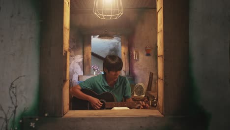 Looking-through-an-open-window-at-a-young-Asian-male-playing-a-guitar-while-on-a-chair-in-a-rustic-room
