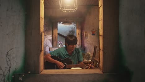 looking through an open window at a young asian male playing a guitar while on a chair in a rustic room