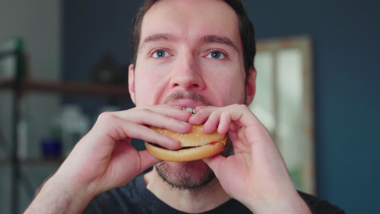 Young caucasian man bites into burger, burger eating pleasure, close up portrait fast food eating, unhealthy eating concept obesity, eating at home alone lonely