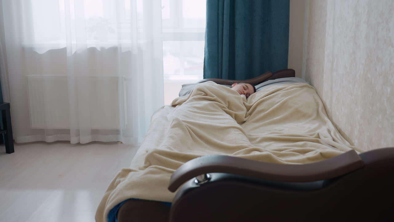 Kid sleeping wrapped in blanket on bed in bright cozy bedroom with soft daylight coming through curtain near window creating calm peaceful atmosphere of rest comfort