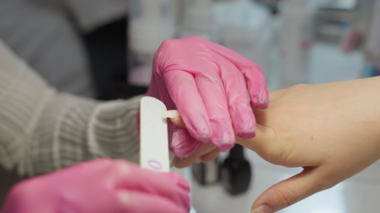Nail technician in pink gloves carefully filing client's nail using emery board, with nail polish bottles softly blurred in background, capturing precise hand care in beauty salon environment