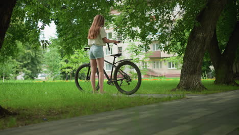 young woman in pink sneakers and jean shorts walks up to her bicycle on a tree-lined path, placing an item on the seat, background includes a building partially visible through the trees