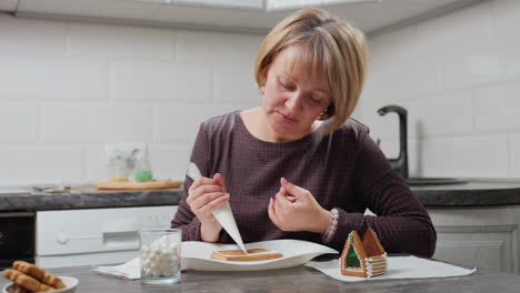 mujer intensamente enfocada en decorar una galleta con glaseado blanco mientras está sentada en una cocina bien iluminada, el fondo presenta galletas borrosas, fregadero de cocina, utensilios y equipos de horneado
