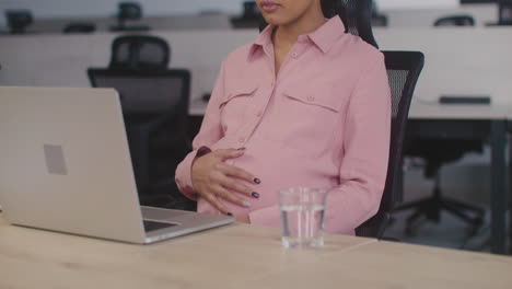 pregnant woman caressing her belly and drinking water while working in the office 1