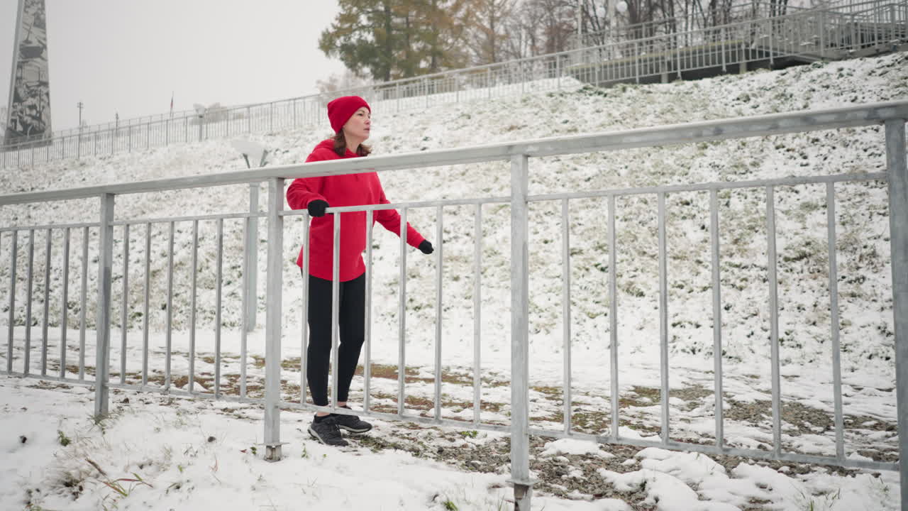 겨울에 야외에서 운동하는 여성, 다리를 밖으로 는 동안 철의 레일링을 들고, 검은 장갑과 빨간 옷을 입고, 눈 인 땅, 안개 대기 및 먼 나무와 함께