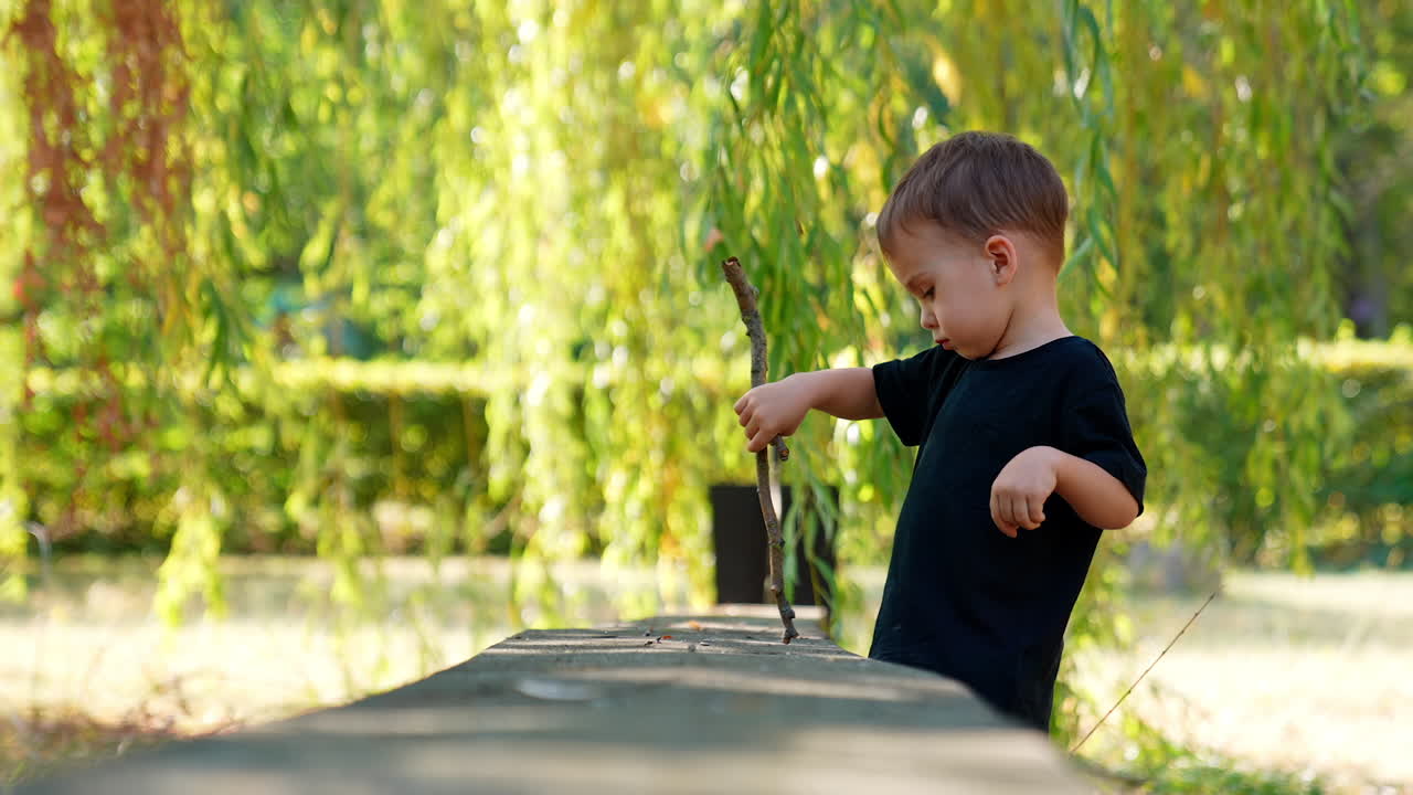 Charming Caucasian baby boy wearing black t-shirt standing under the willow tree. Kid holds a stick and taps with it by the surface.