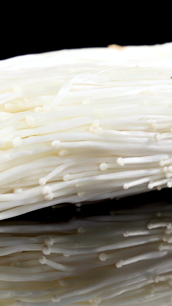 A bundle of enoki mushrooms slowly rotates on a reflective black surface, illuminated by soft studio lighting, highlighting their delicate texture and white color