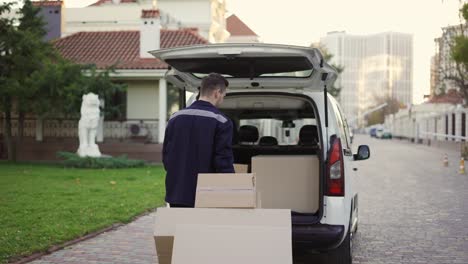 joven apuesto hombre de entrega caucásico sonriente con cajas en el carro moviéndose hacia la furgoneta con el camión abierto. trabajador masculino de la oficina de correos