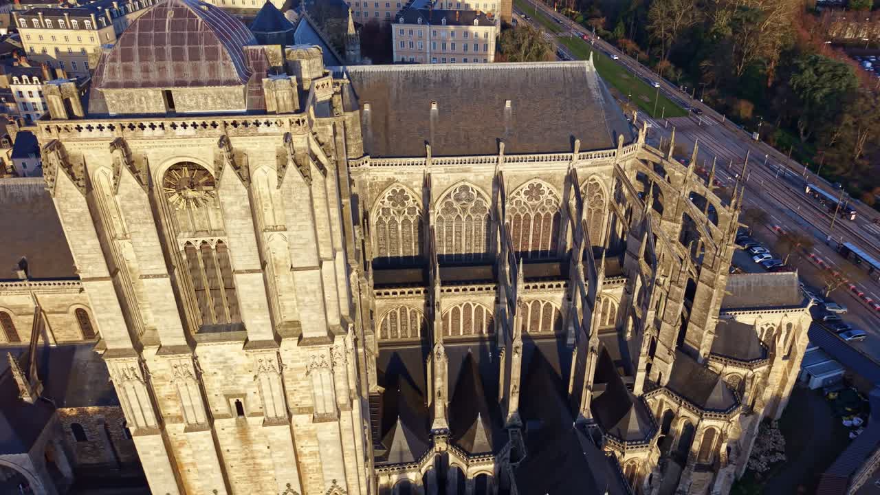 Drone orbitando a torre principal da Catedral de Le Mans ao pôr do sol, com a luz dourada aquecendo a fachada de pedra enquanto a paisagem urbana circundante e as ruas aparecem abaixo. França.