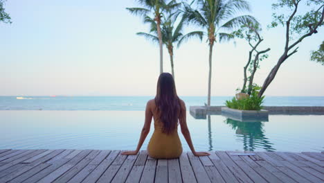 locked-medium-wide-silhouette-shot-of-young-adult-Asian-woman-in-swimsuit-sitting-at-the-edge-of-infinity-pool-watching-tropical-magic-hour-on-the-ocean-with-palm-trees-and-copy-space-no-face