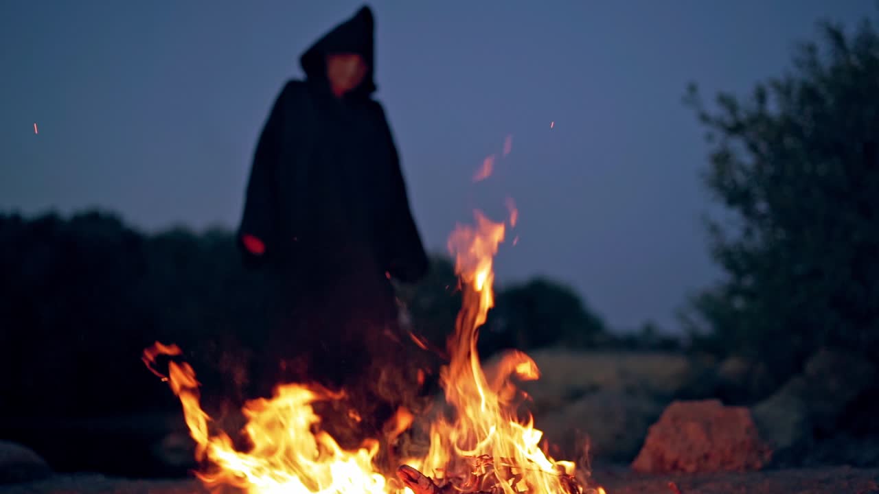 Bright fire flame and black witch behind. Spooky figure in black cloak standing near the burning fire at night. Halloween concept.